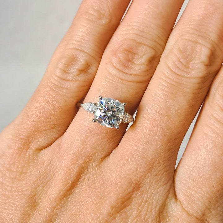 Cushion and pear cut moissanite three stone engagement ring in white gold, close up on ring finger