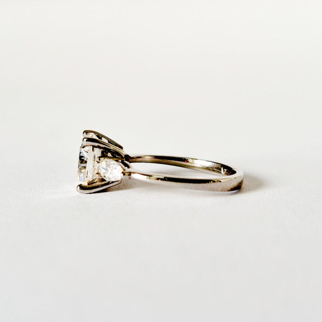 Cushion and pear cut moissanite three stone engagement ring in white gold, side view on white background