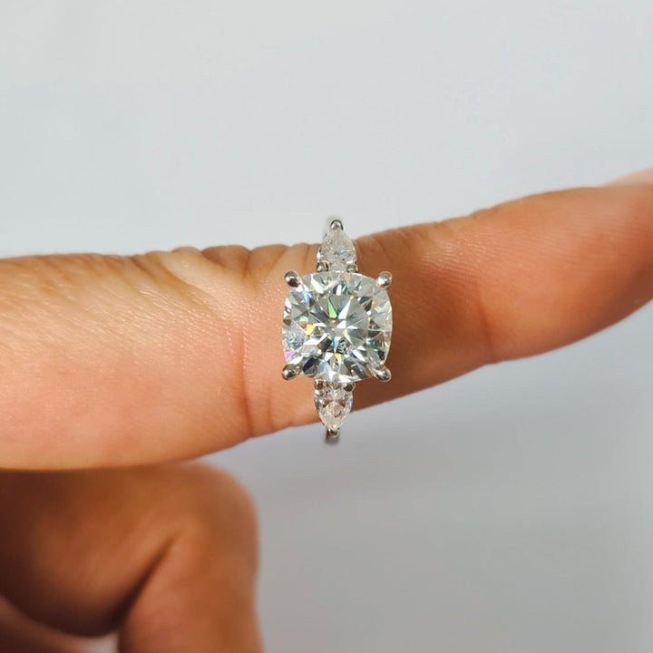 Cushion and pear cut moissanite three stone engagement ring in white gold, close up on finger