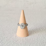 1.5 carat oval halo moissanite engagement ring set in platinum next to eternity wedding band seen on wooden cone and grey background